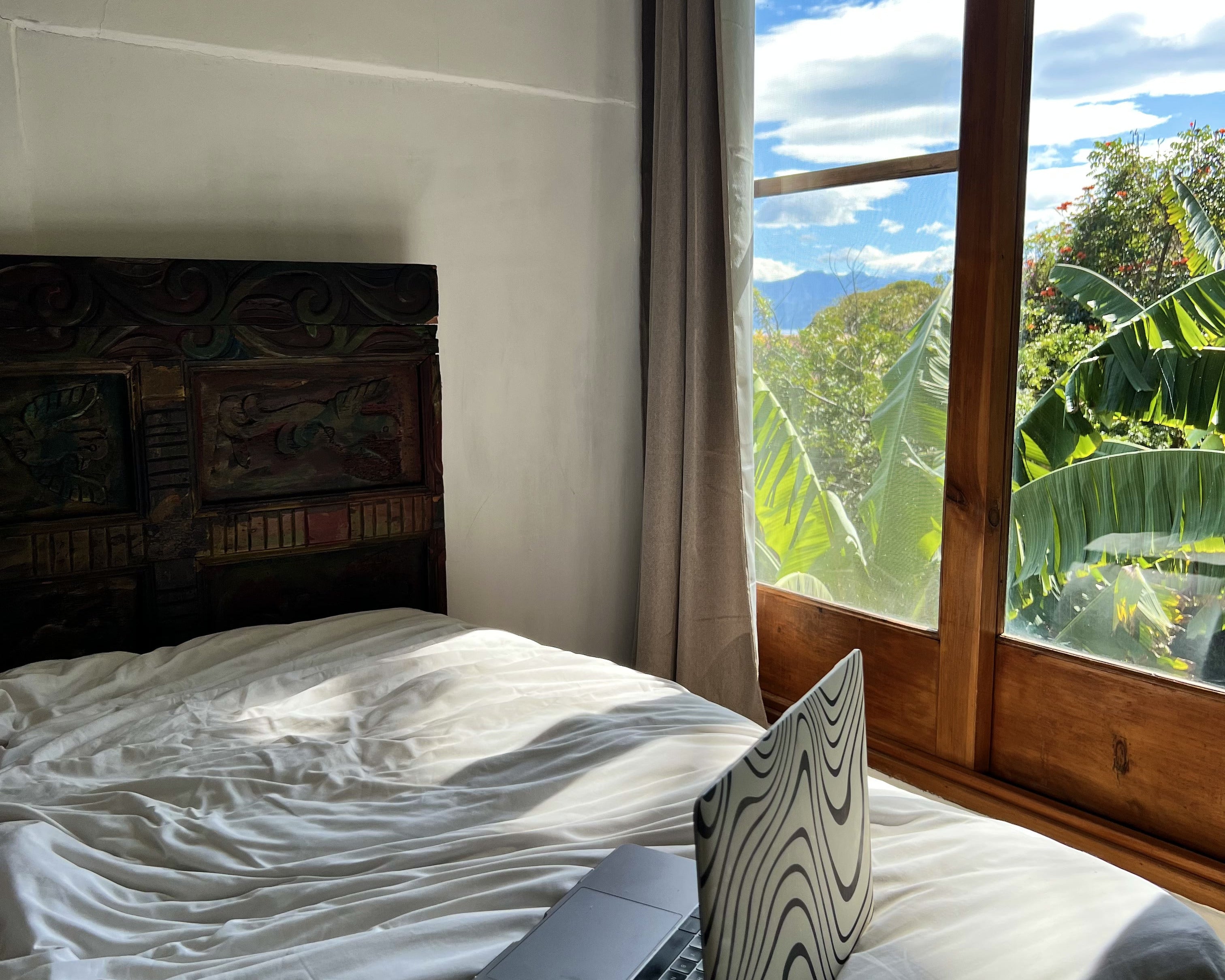 Laptop on a bed with a large window showing a scenic view of trees and blue sky.